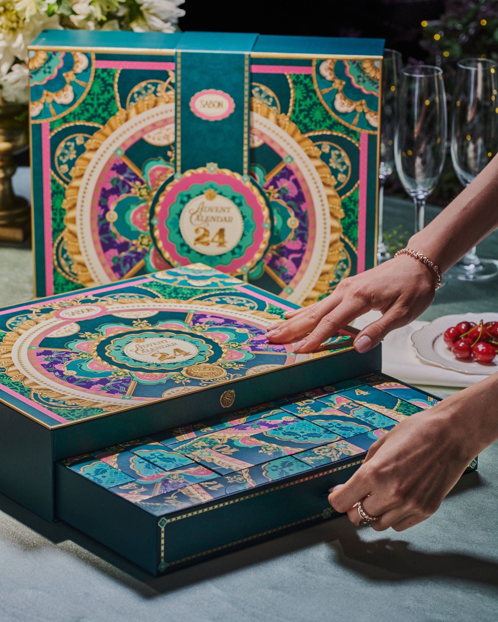 Decorative advent calendar with colorful design, being opened by a hand on a table with glasses and flowers.